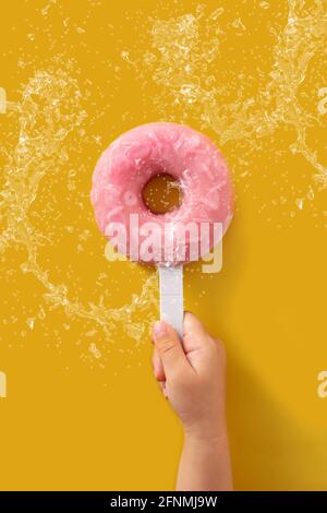 Hand hält einen Erdbeerdonut Popsicle spritzt Wasser auf gelb Hintergrund Stockfoto