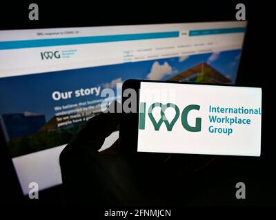 Person, die das Smartphone mit dem Logo des multinationalen Serviced Offices-Unternehmens IWG plc auf dem Bildschirm vor der Website hält. Konzentrieren Sie sich auf die Telefonanzeige. Stockfoto