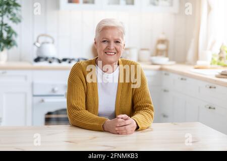 Lächelnde ältere Dame, die zu Hause am Tisch in der Küche sitzt Stockfoto