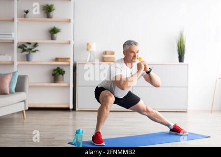 Workout zu Hause. Reifer Mann trainiert und macht seitliche Ausfallschritte auf der Matte im Wohnzimmer Innenraum zu Hause, leerer Raum Stockfoto