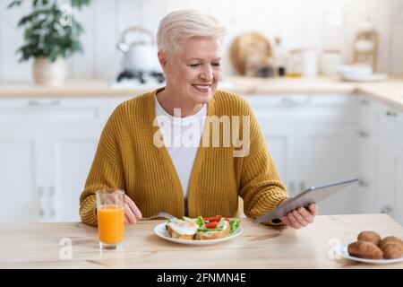 Positive ältere Frau, die zu Mittag gegessen und ein digitales Tablet benutzt Stockfoto
