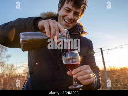 Porträt eines aufgeregten Mannes, der Rotwein aus dem Krug ausgießt Im Winter Glas vor dem Weinberg Stockfoto