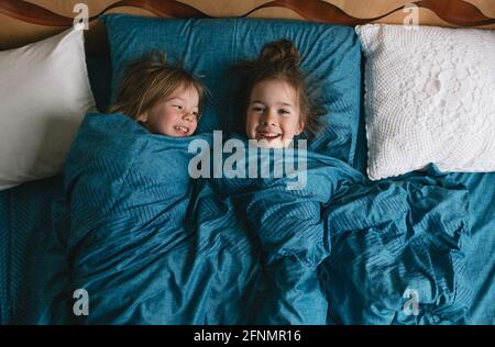 Von oben schauen zwei entzückende Töchter unter der Decke, schauen in die Kamera, liegen zusammen auf dem gemütlichen Bett zu Hause, nette Kinder entspannen sich, genießen Stockfoto