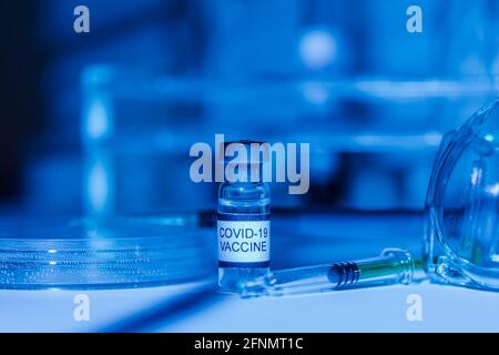 Labortisch mit Halterung für Reagenzgläser, Fläschchen mit Impfstoff für COVID-19, Spritze, Pipette, Schutzbrille und Petrischale, mit blauem Licht Stockfoto