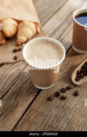 Zwei Pappbecher Kaffee, Croissants bündeln sich am Sacktuch, getrocknete geröstete Kaffeebohnen auf Löffel am alten Holztisch Stockfoto