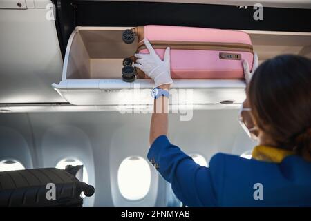 Mitarbeiter der Fluggesellschaft versteckt Handgepäck auf Rädern im Gepäckraum über dem Kopf Stockfoto