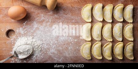 Hausgemachte Eiernudeln. Ungekochte gefüllte Ravioli mit Ricotta und Spinat auf dem Pappbrett, mit Mehl, Ei und Nudelholz. Draufsicht, Platz für Text. Stockfoto