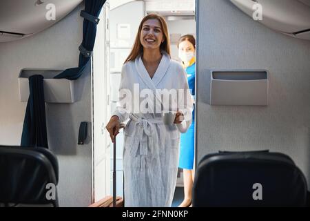 Entspannte Frau, die in der Flugzeugkabine mit Bademantel und Kaffee kommt Stockfoto