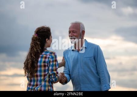 Älterer Mann und junge Frau schütteln sich im Freien mit Dramatic die Hände Himmel bei Sonnenuntergang im Hintergrund Stockfoto