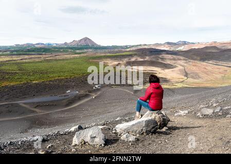 Frau, die auf einem Felsen auf einem sitzt Schlafender Vulkan mit Blick auf eine dramatische Vulkanlandschaft in Island An einem bewölkten Sommertag Stockfoto