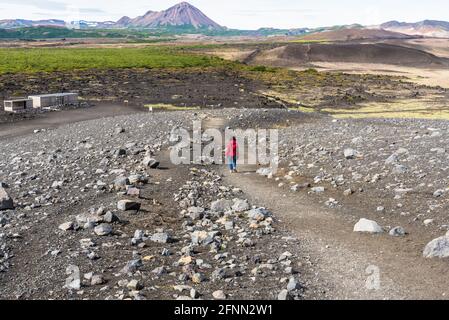 Einsame Wanderin roter Jacke gehende Frau, die einen Steilhang hinuntergeht Pfad entlang der Seite eines Dorment Vulkans in Island An einem bewölkten Sommertag Stockfoto