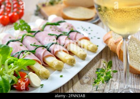 Köstlicher Frühlingssalat mit Spargel, gerollt in gekochtem Schinken auf einer weißen Platte, serviert mit einem jungen trockenen Weißwein und Baguette-Scheiben Stockfoto