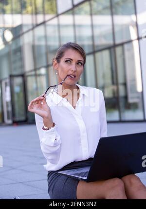 Nachdenkliche, hübsche junge Geschäftsfrau, die auf der Treppe vor dem Bürogebäude sitzt, eine Brille hält und im Schoß am Laptop arbeitet Stockfoto