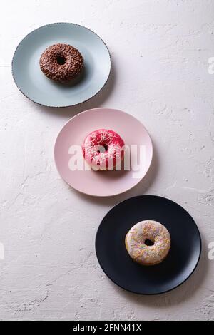 Drei Donuts in Reihe auf Tellern, Chocolate-, Pink- und Vanilledonut mit Streuseln, süß glasierte Dessertnahrung auf weißem Beton strukturiertem Hintergrund, Angl Stockfoto