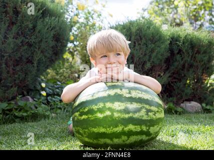 An einem sonnigen Sommertag sitzt ein süßes blondes Kleinkind auf den Knien auf dem leuchtend grünen Gras in der Nähe einer großen Wassermelone und lächelt süß. Warten auf ein Stockfoto