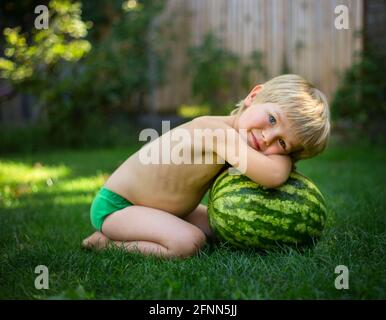 An einem heißen Sommertag sitzt ein süßer barfuß kleiner Junge ohne Kleidung auf dem Gras neben einer großen gestreiften Wassermelone. Warten auf einen appetitlichen Feinschmeckergeschmack Stockfoto