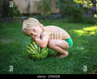 An einem heißen Sommertag sitzt ein süßer barfuß-Kleinkind neben einer großen gestreiften Wassermelone. Warten auf ein köstliches Dessert. Fröhliche, unbeschwerte Kindheit Stockfoto