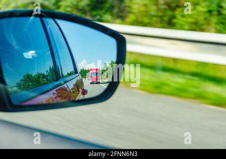 Spiegelung eines verschwommenen roten LKWs im Rückspiegel an autobahn in schönen sonnigen Tag Stockfoto