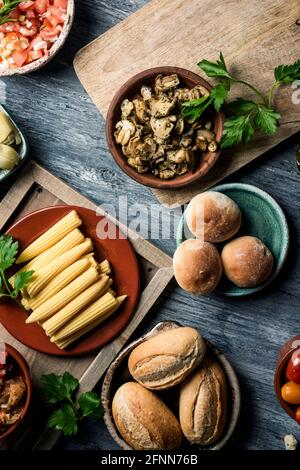 Blick aus der Perspektive auf einige verschiedene Zutaten für die Zubereitung veganer Sandwiches, wie z. B. Brotbrötchen, gekochte Pilze, Baby-Mais, gehackte Tomaten und Lch Stockfoto