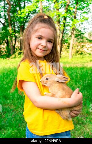 Ein kleines süßes Mädchen in einem gelben T-Shirt hält ein echtes Kaninchen in den Armen vor einem Hintergrund grüner Pflanzen. Im Sommer Aktivitäten im Freien für Kinder Stockfoto