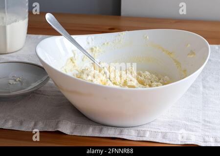 Quark-Teig in einer weißen Schüssel mit einer Gabel auf einem Leinentischtuch auf einem Holztisch mischen. Zutaten - Quark, Butter, Ei und Zucker. Stockfoto