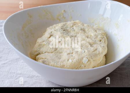 Roher Quark-Teig mit Stückchen Quark in einer weißen Schüssel auf einem Leinentischtuch auf einem Holztisch. Der Prozess der Herstellung von Quark Bagels oder Croissants Stockfoto