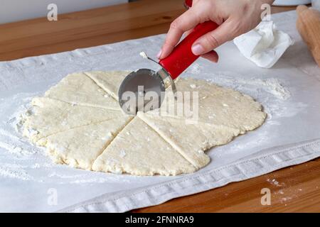 Auf einem Leinentischtuch auf einem Holztisch schneidet man gerollten rohen Quark-Teig mit einem runden Pizzabmesser von Hand in acht Dreiecke auf Backpapier. Der Prozess von Stockfoto