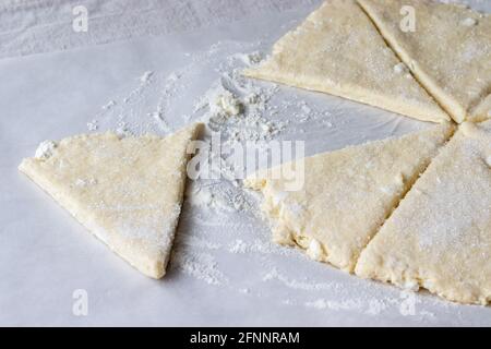 Gerollter roher Quark, der in acht mit Zucker bestreute Dreiecke geschnitten wird, und ein Teigdreieck liegt getrennt auf Backpapier auf einer Leinentischdecke. T Stockfoto