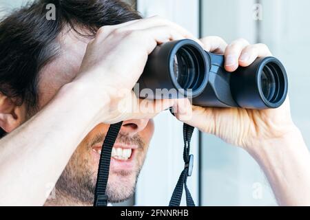 Ein gutaussehender, lächelnder bärtiger Mann, der durch ein Fernglas auf das Fenster blickt und Blick auf das Nachbarhaus hat, um Nachbarn, Umgebung oder Nat zu beobachten Stockfoto