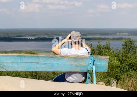 Ein Mann blickt durch ein Fernglas auf einen malerischen Blick auf einen breiten Fluss, während er auf einer alten Holzbank auf einem Hügel sitzt. Rückansicht Stockfoto