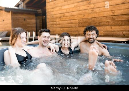 Gesellschaft von Freunden dampft im Whirlpool mit Blasen im Freien in der Nähe des Spa und See. Frauen und Männer haben Spaß und entspannen sich im Wellnesscenter. Stockfoto