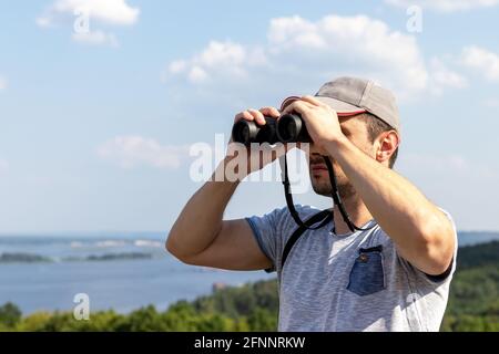 Ein Mann schaut durch ein Fernglas auf eine landschaftlich reizvolle Aussicht Ein breiter Fluss auf einem Hügel an einem sonnigen Tag Stockfoto