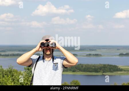 Ein Mann, der durch ein Fernglas die Kamera anschaut, gegen eine Szenenbildkamera Blick auf einen breiten Fluss auf einem Hügel auf einem Sonniger Tag Stockfoto