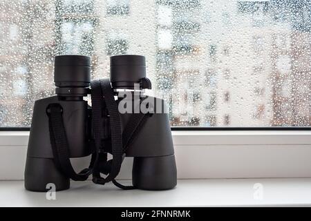 Auf einer weißen Fensterbank am Fenster mit Regentropfen auf dem Glas befindet sich ein schwarzes Fernglas zur Beobachtung von Nachbarn. Faszinierende Beobachtung der Umwelt Stockfoto