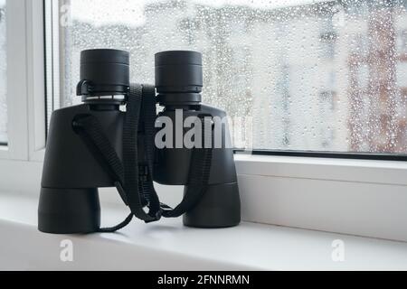 Schwarzes Fernglas zur Beobachtung von Nachbarn befindet sich auf einer weißen Fensterbank am Fenster mit Regentropfen auf dem Glas. Faszinierende Beobachtung der Umgebung Stockfoto