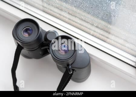 Nahaufnahme von oben auf einem schwarzen Fernglas zur Beobachtung von Nachbarn oder der Natur, das auf einer weißen Fensterbank am Fenster mit Regentropfen auf dem Glas steht. Stockfoto