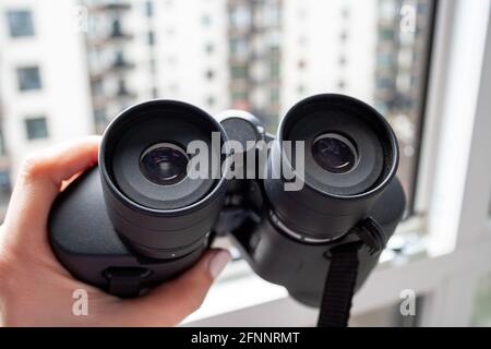 Nahaufnahme einer weiblichen Hand, die ein schwarzes Fernglas gegen sich hält Ein verschwommener Hintergrund eines Fensters mit Blick auf Das Nachbarhaus für die Beobachtung nei Stockfoto