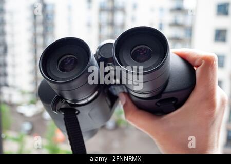 Hand hält ein schwarzes Fernglas vor einem verschwommenen Hintergrund eines Fensters mit Blick auf das Nachbarhaus, um Nachbarn, Umgebung oder na zu beobachten Stockfoto