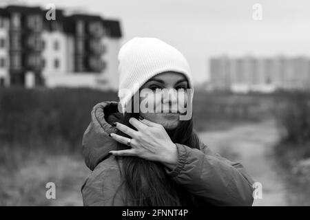 Lächelnd schöne junge Frau mit langen Haaren in gestricktem Hut auf der Straße an einem bewölkten Tag vor verschwommenem Hintergrund. Weibchen ohne Make-up, natürlich Stockfoto