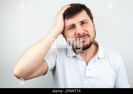 Porträt eines hübschen bärtigen Mannes mit verengten Augen und Lippen, der seinen Kopf mit der Hand hält und unter Kopfschmerzen leidet, in einem weißen T-Shirt auf einem WH Stockfoto