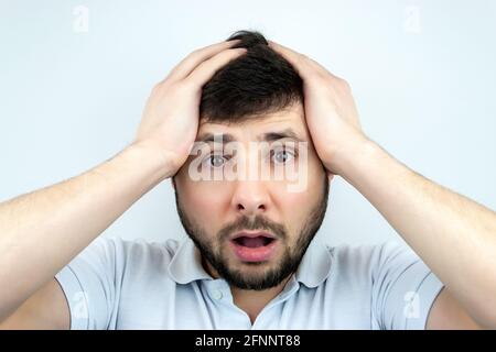 Überraschender bärtiger Mann mit weit geöffneten Augen und Mund, der seinen Kopf mit den Händen in einem weißen T-Shirt auf weißem Hintergrund hielt Stockfoto