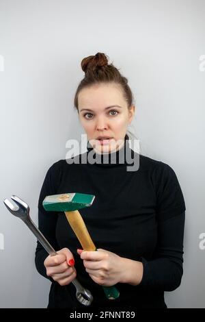 Überraschte Frau mit gebrümmten Haaren und schwarzem Pullover mit einem Hammer und ein großer Schraubenschlüssel in ihren Händen Stockfoto