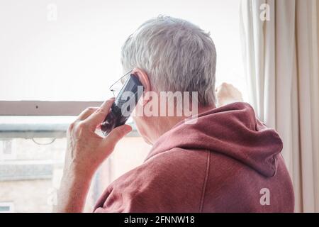 Älterer grauhaariger Mann, der das Telefon benutzte und aus einem Fenster schaute, London, Großbritannien Stockfoto