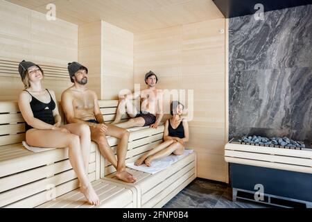 Gesellschaft von vier Freunden, die sich in heißer traditioneller Sauna in Saunahüten entspannen. Wellness und Freizeit im Spa. Stilvolles Interieur mit Tageslicht. Stockfoto