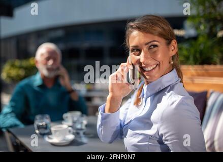 Zwei Geschäftsleute, ein älterer Mann und eine junge Frau, sitzen im Café, unterhalten sich mit dem Handy und warten auf jemanden Stockfoto