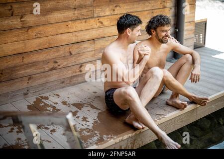 Männliche Freunde sitzen auf der Terrasse in der Nähe des Sees im Spa-Komplex, während sie reden und die Hände halten. Entspannen Sie sich und genießen Sie die Zeit im Wellness-Spa. Stockfoto