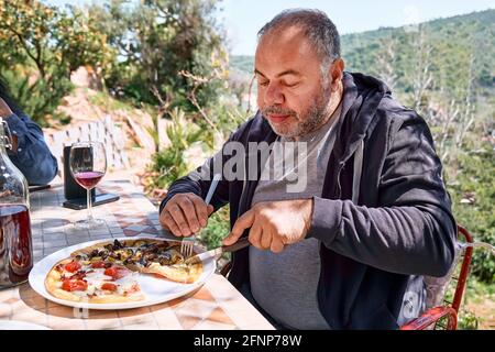 Mann, der Pizza auf dem Teller auf dem Kacheltisch im Garten zerschnitt. Hausgemachte italienische Pizza mit Tomaten, Käse und Schinken. Stockfoto
