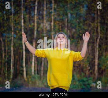 Ein junges Mädchen mit langen Haaren geht mit erhobenen Armen durch den Wald. Blonde Haare. Gelbe Jacke. Stockfoto