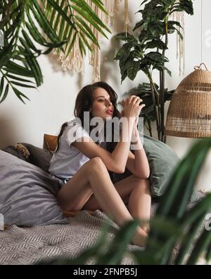 Die attraktive junge Frau entspannt sich auf einem bequemen Bett in einem gemütlichen Schlafzimmer. Boho Chic Interieur Stil mit heimischen Pflanzen und Macrame Stockfoto