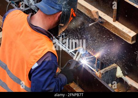 Ein Schweißer in einem Schutzhelm und Handschuhen arbeitet mit Metall, schweißt Metallstäbe während des Baus einer Holzkiste zum Gießen einer Betonkonstruktion Stockfoto